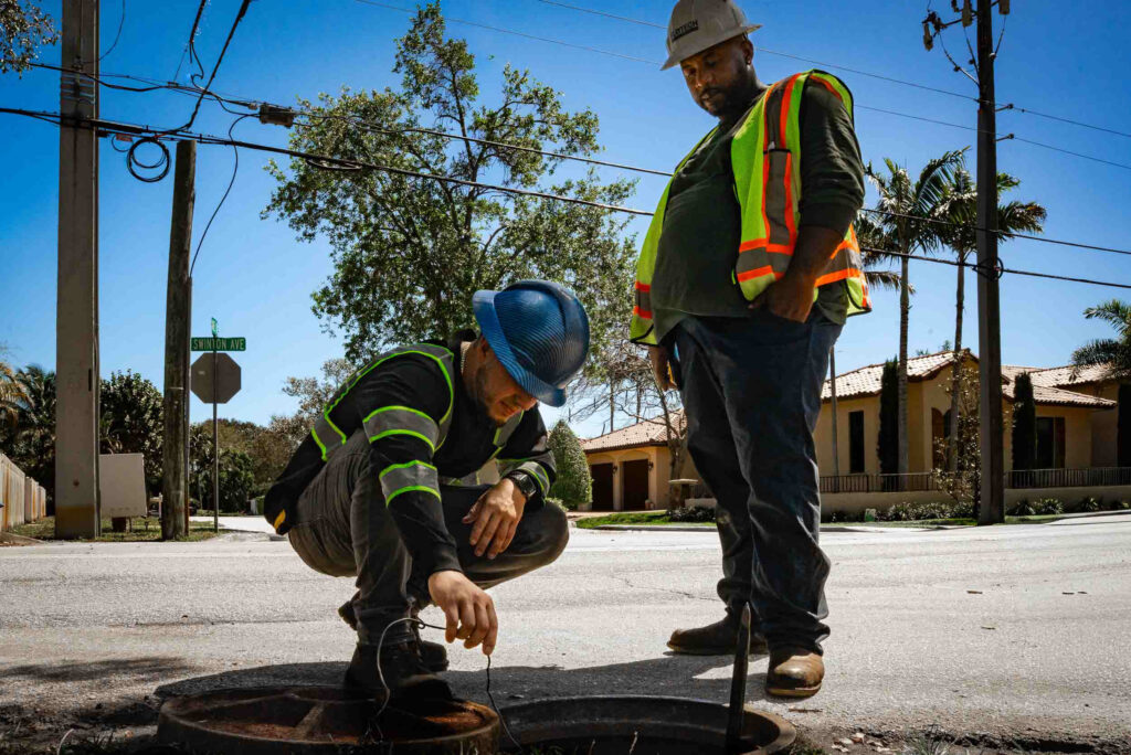 Worker conducting a manhole inspection, wearing safety gear and using specialized equipment on a city street.